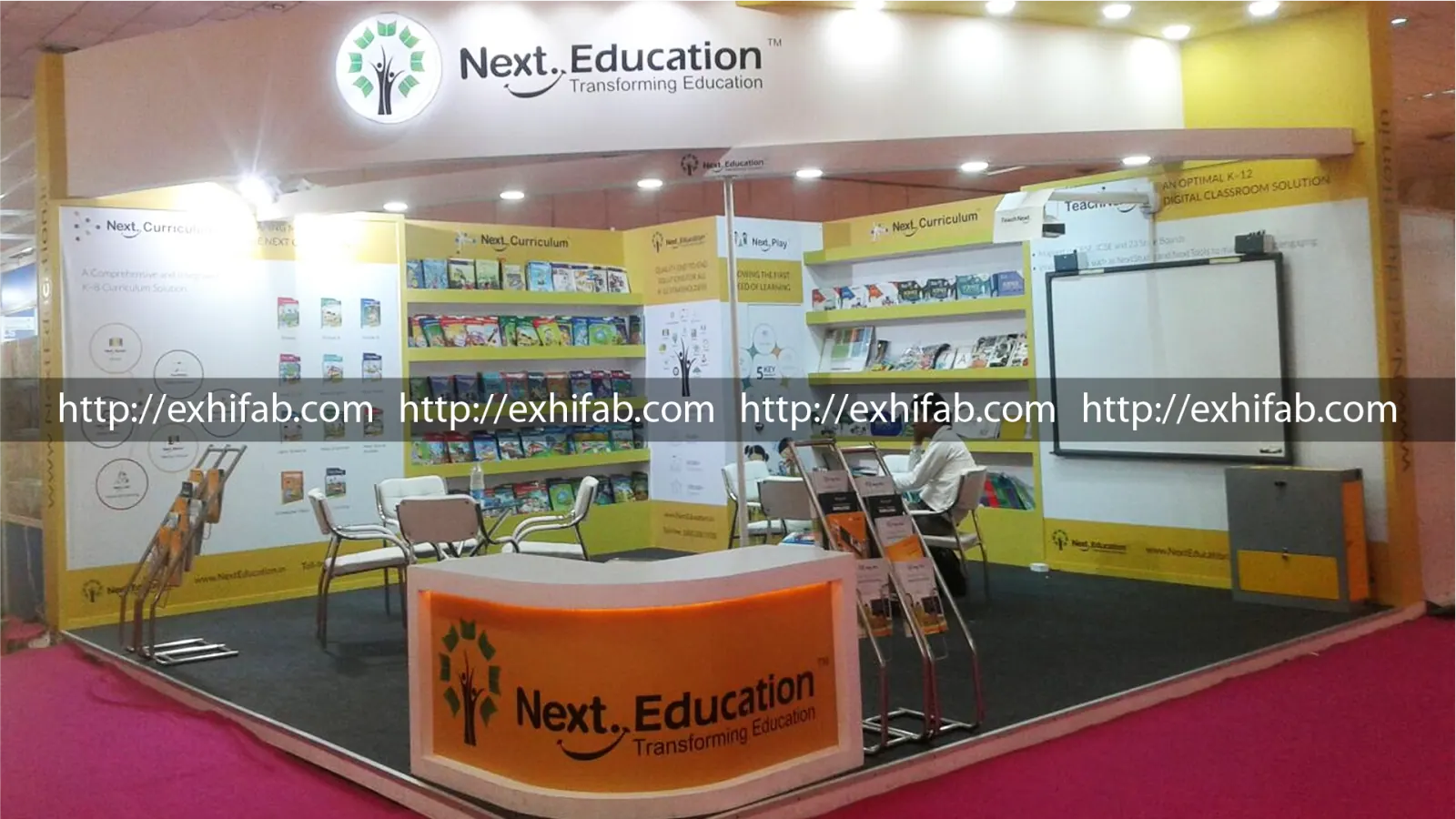 Next Education exhibition stall with backlit logo, tiered book display shelving, curriculum panels and interactive smartboard